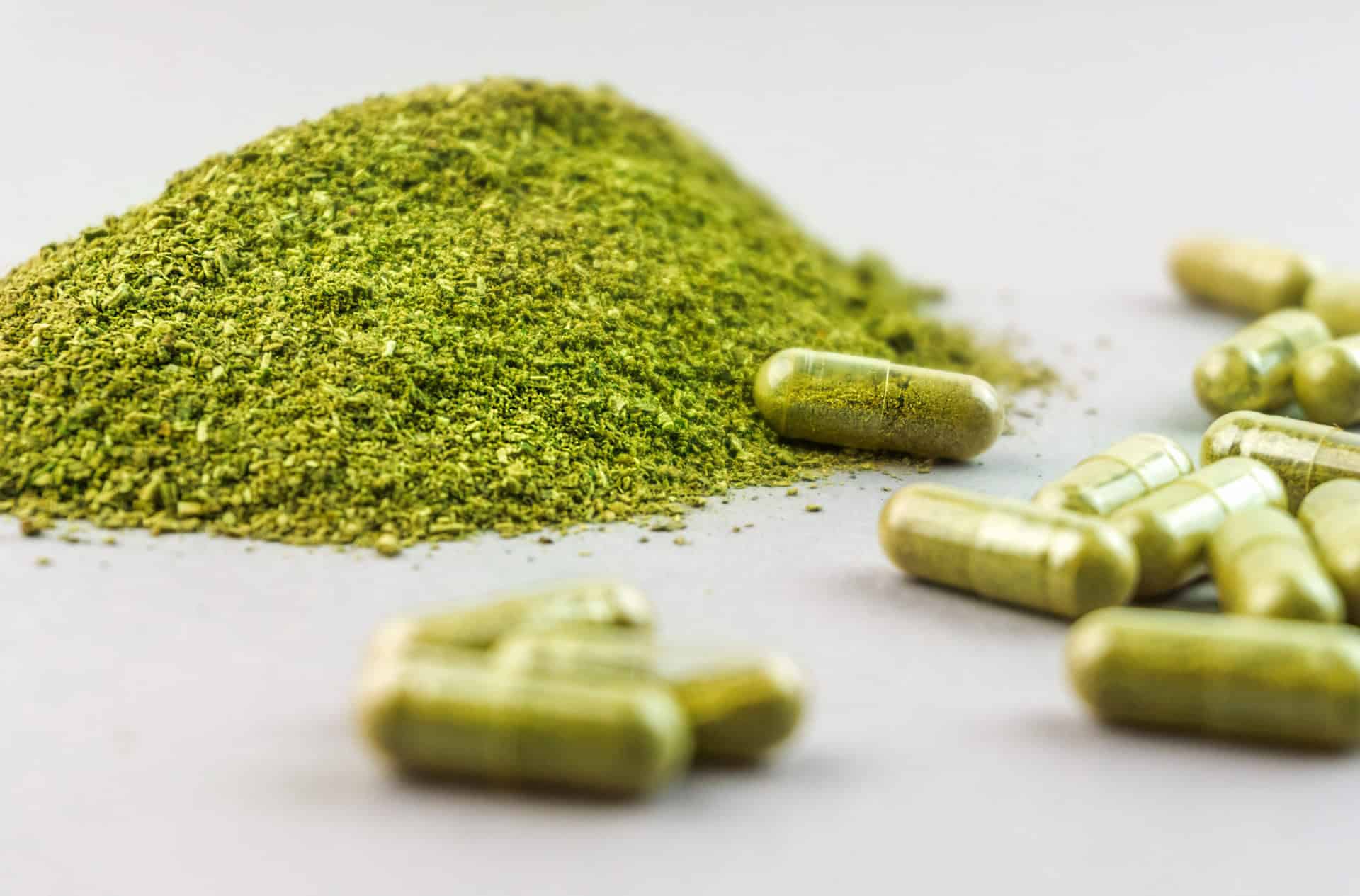 A pile of green kratom powder alongside several filled capsules on a white surface, providing a clear visual of the substance in its common forms.