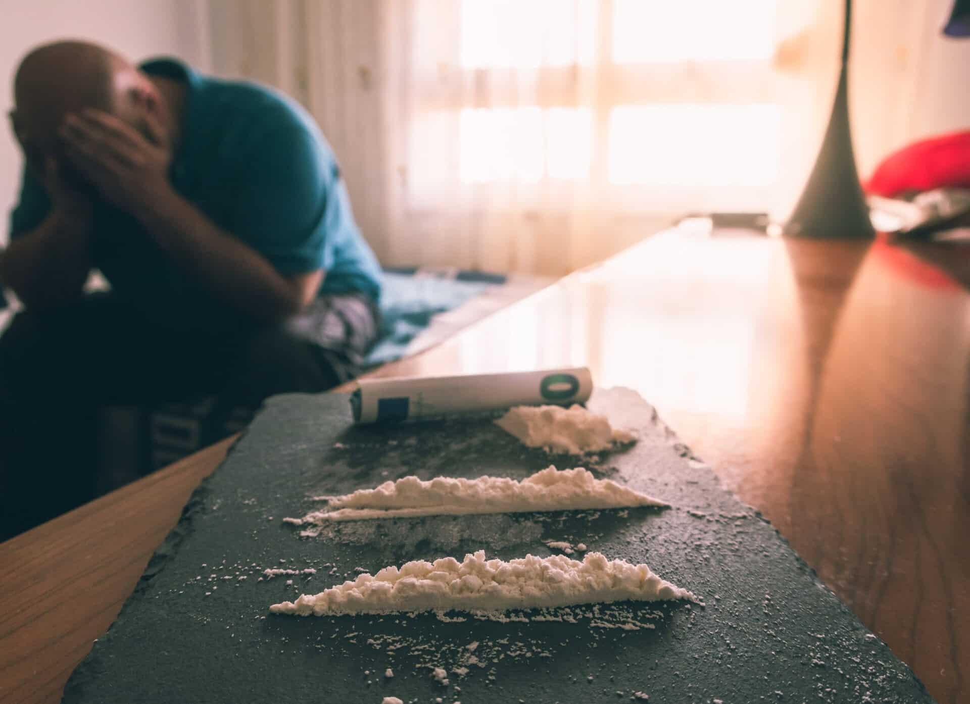 Close-up of cocaine lines on a dark surface with a blurred man in the background holding his head in distress, illustrating the physical and emotional impact of stimulant use.