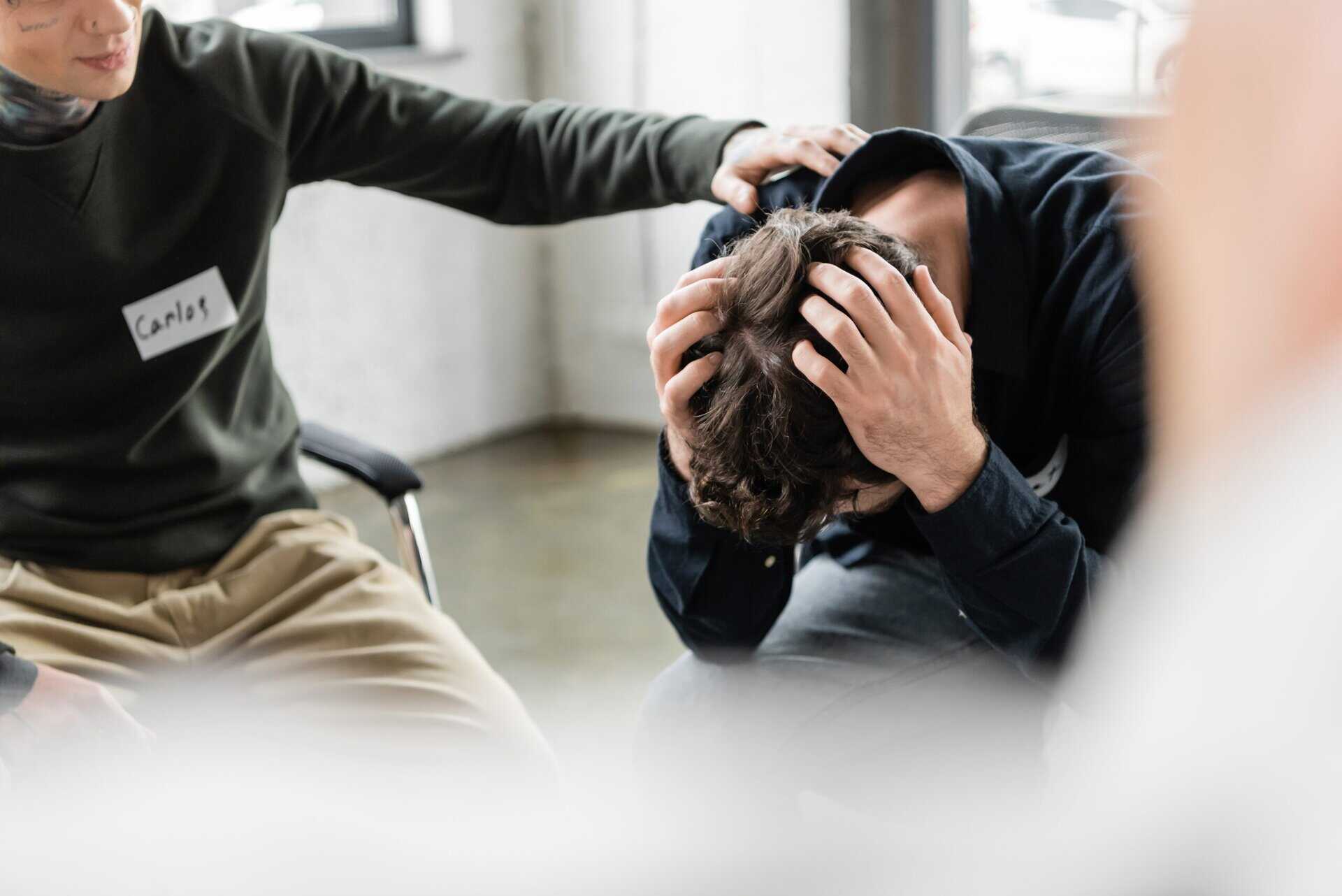 An emotional scene in a support group where a man sits with his head in his hands while a peer or counselor places a supportive hand on his shoulder.