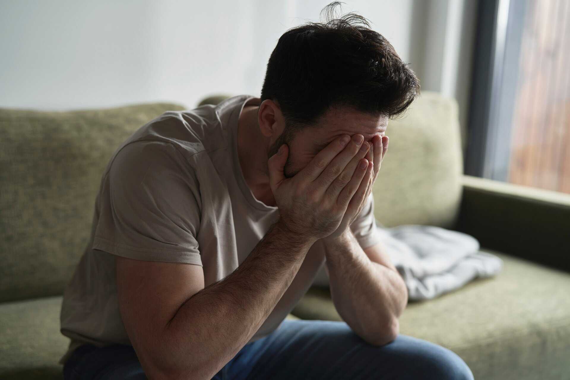 A man sitting on a sofa with his head in his hands, depicting the isolation, depression, and 'crash' phase associated with stimulant withdrawal.