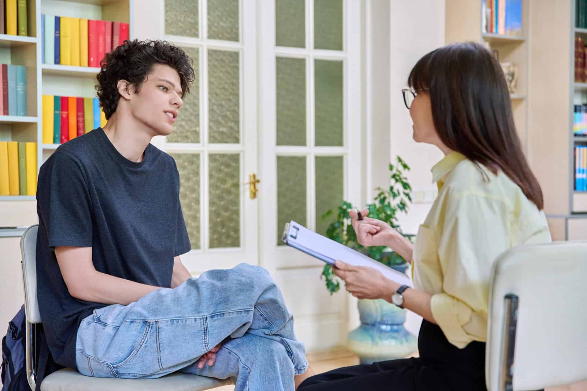 A focused one-on-one therapy session between a young male patient and a female clinical psychologist in a modern office with bookshelves in the background.