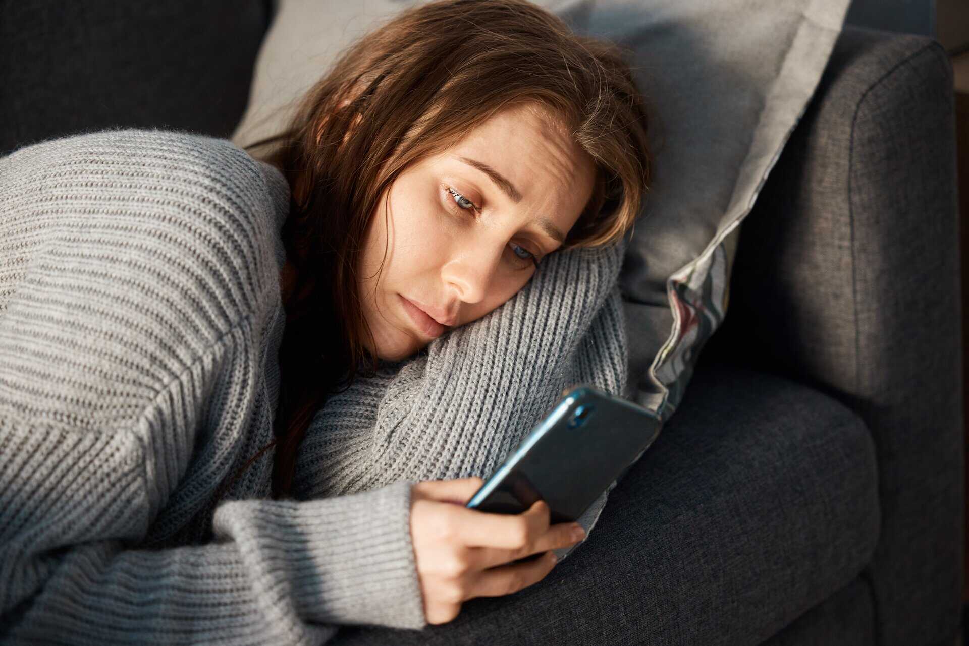 A woman lying in bed looking at her phone with a somber expression, depicting the emotional toll and loneliness of struggling with dependency.