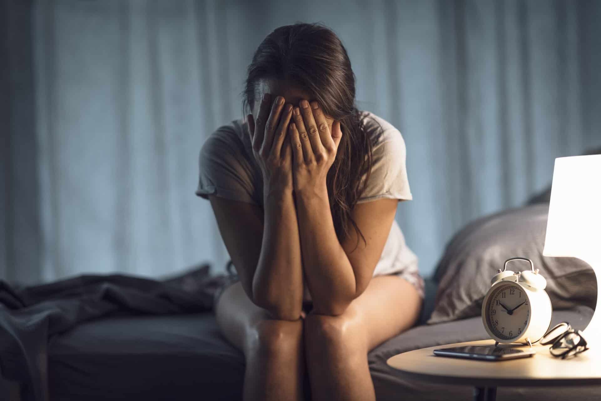 A woman sitting on a bed in a dark room with her face buried in her hands.